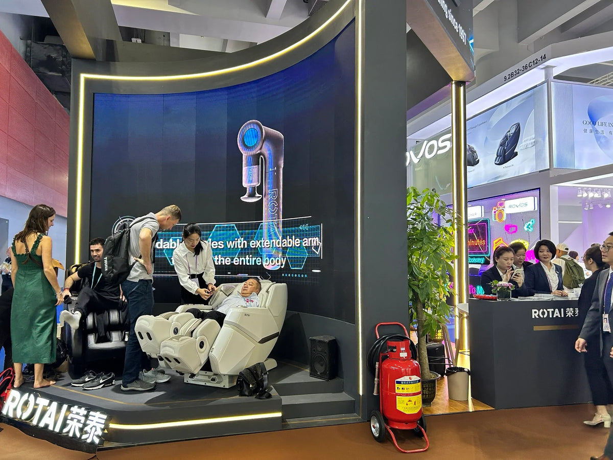 Expo booth with people trying ROTAI massage chairs, large digital display, and staff assisting.